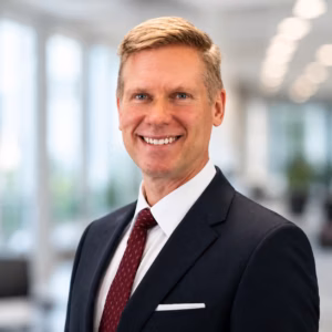 Professional head-and-shoulders portrait of a smiling man in a navy suit and red tie in a bright office setting.
