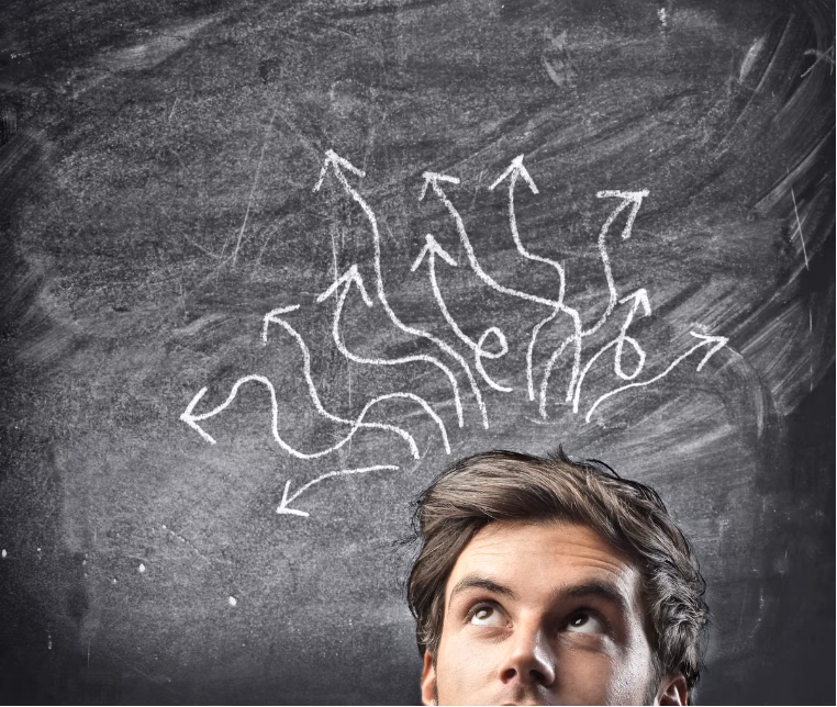 Man looking up at a chalkboard filled with many tangled arrows and curves, suggesting ideas or decision paths