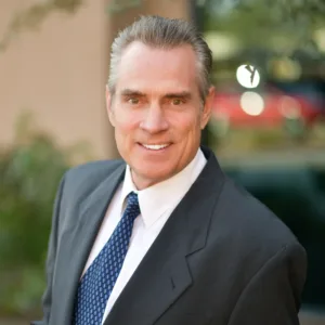 Smiling man in a dark suit and blue tie outdoors, facing slightly to the right with a blurred natural background